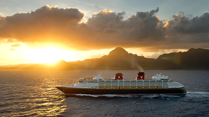 Un barco de Disney Cruise Line pasa navegando por montañas donde se está poniendo el sol